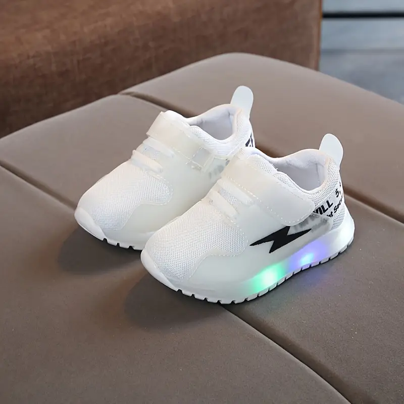 Weiße LED-Kinderschuhe mit Klettverschluss auf grauem Sofa, leuchtende Sohle.