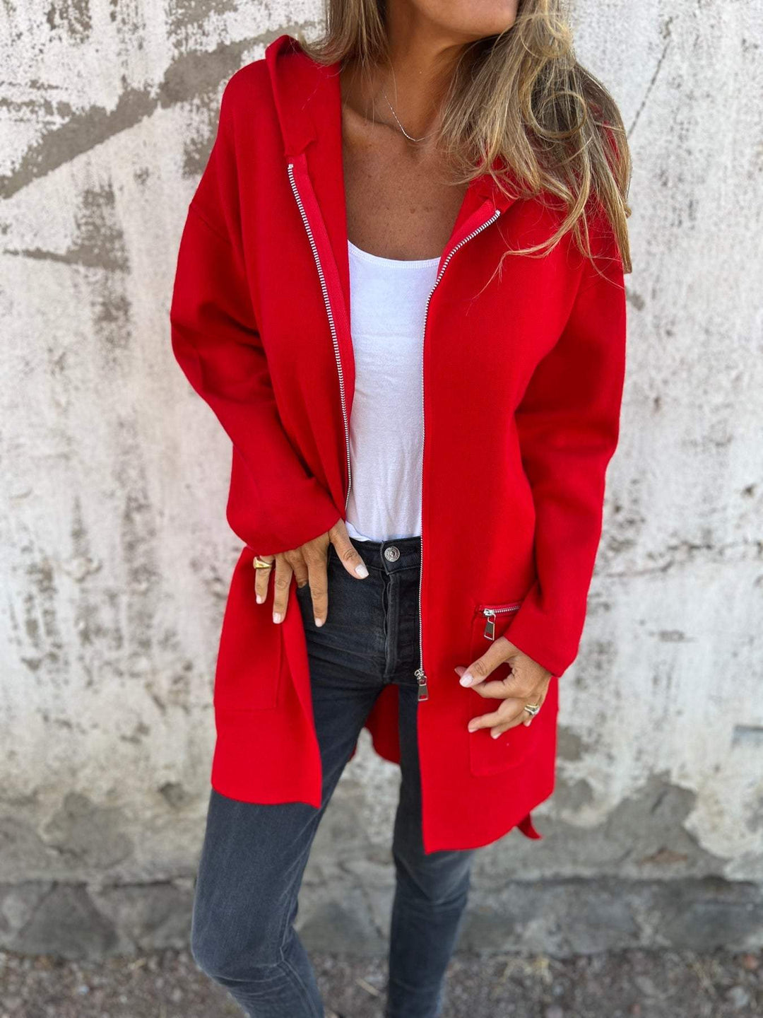 Frau in roter Kapuzenjacke mit Reißverschluss, weißem Top und schwarzer Jeans vor Betonwand.