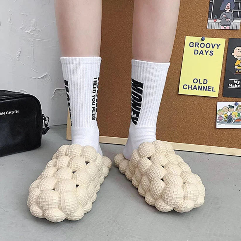 Beige 3D-Bubble-Slipper aus Kunststoff, getragen mit weißen Socken, modisch und bequem.