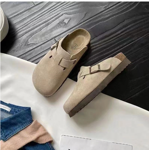 Graue Wildleder-Schlupfclogs mit Schnalle auf dunklem Holzboden, modische bequeme Schuhe.