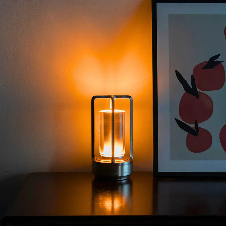 Moderne Tischlampe aus Metall mit warmem Licht neben Kunstbild auf dunklem Tisch.