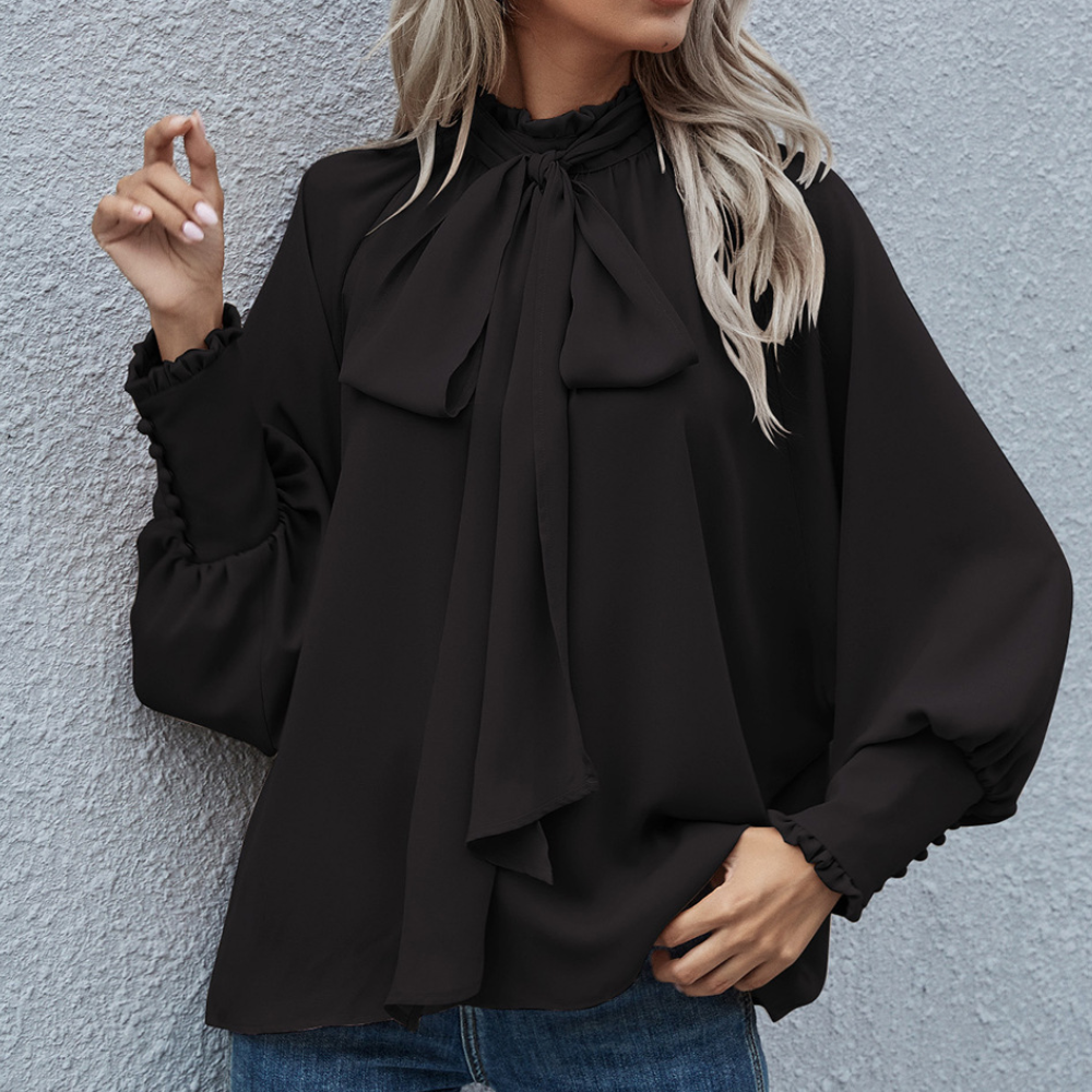 Frau in schwarzer Bluse mit Schleife, lange Ärmel, elegante Damenmode, vor grauer Wand.