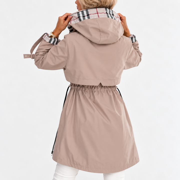 Béžový kabát zezadu na modelce, která drží okraje kapuce s kostkovanou podšívkou, vzadu je vidět stažení v pase šňůrkou. 3