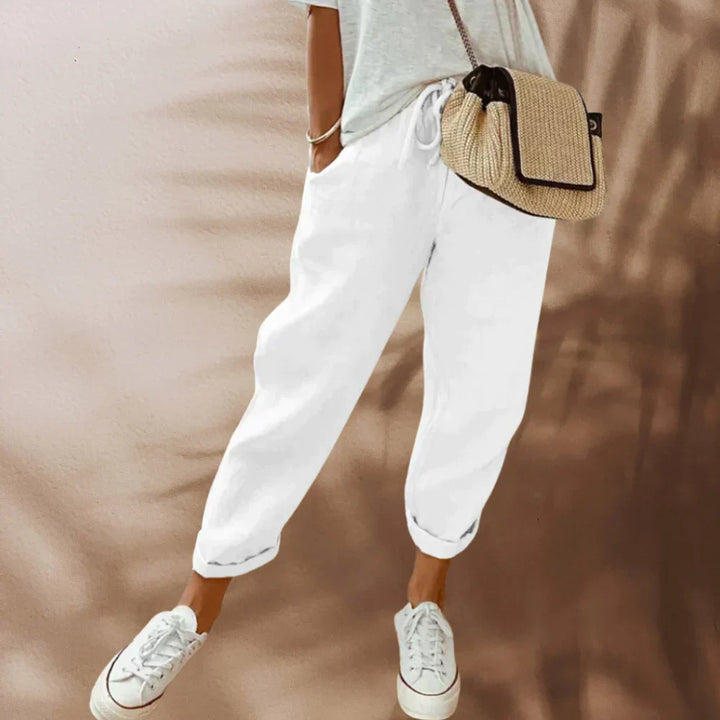 Frau in weißer Leinenhose, Sneakers und Strohtasche vor beigem Hintergrund.