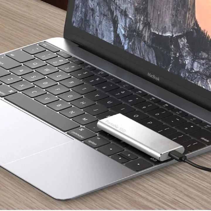 Stříbrný externí disk připojený kabelem k notebooku, položený přímo na jeho klávesnici na dřevěném stole. 7