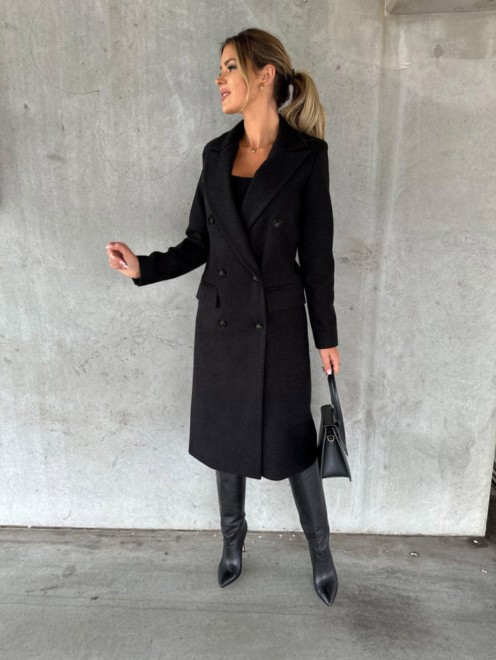 Frau im eleganten schwarzen Wollmantel, trägt Stiefel und Handtasche, vor Betonwand.