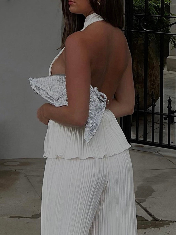 Frau in weißem, rückenfreiem Jumpsuit mit Plissee-Design und silberner Clutch.