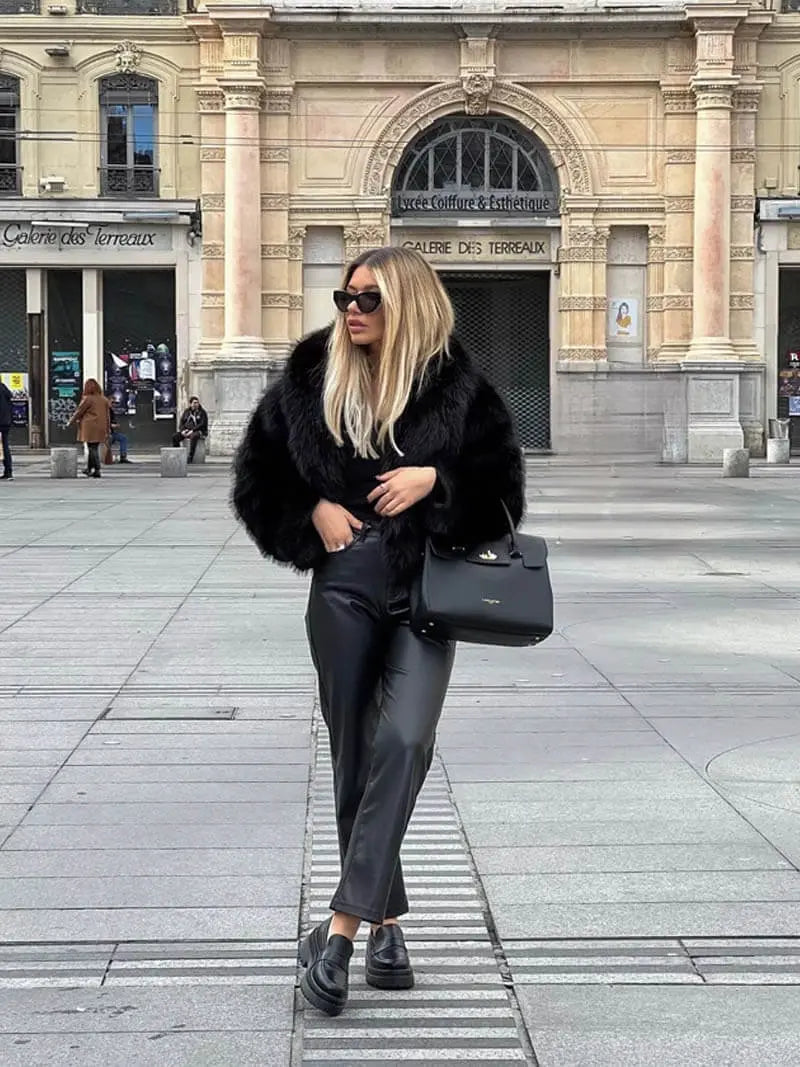 Modelka v černém kožešinovém kabátě, koženkových kalhotách a masivních mokasínech stojí na dlážděném náměstí 8
