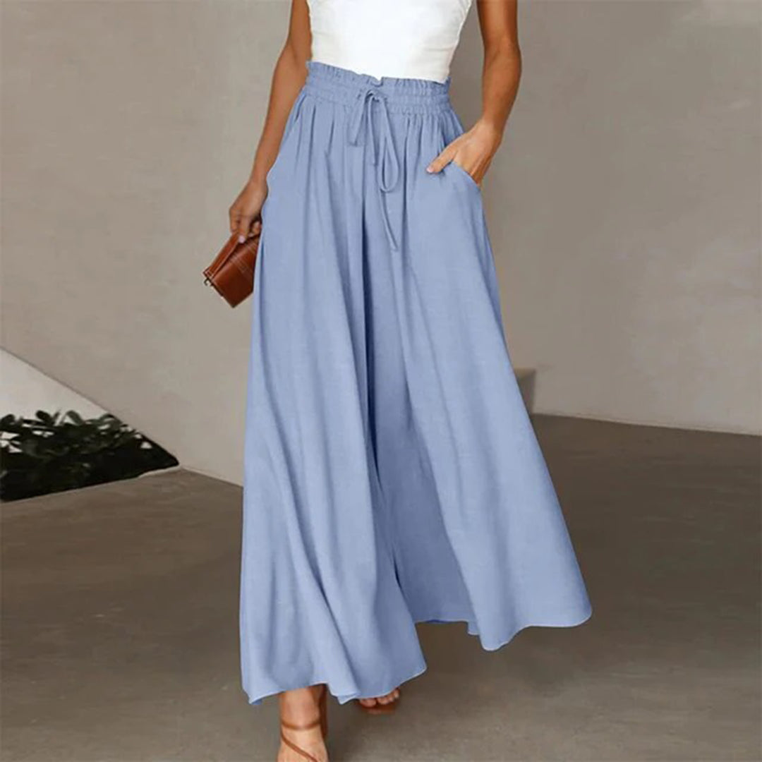 Frau in blauer, weiter Hose aus Leinen, mit weißem Top und brauner Tasche, modisch elegant.