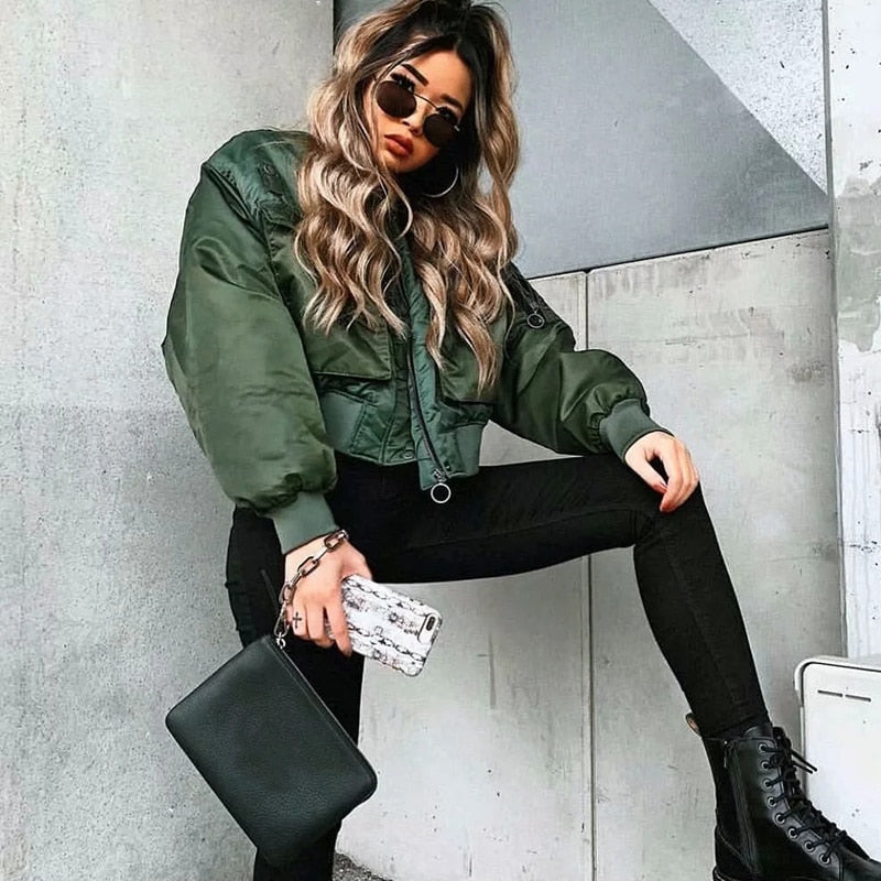 Frau in grüner Bomberjacke, schwarze Hose, Sonnenbrille, mit Handtasche und Smartphone.