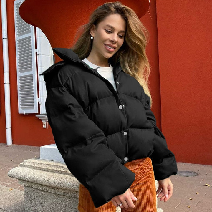 Frau in schwarzer Daunenjacke, lächelnd, vor roter Wand, modische Winterbekleidung.