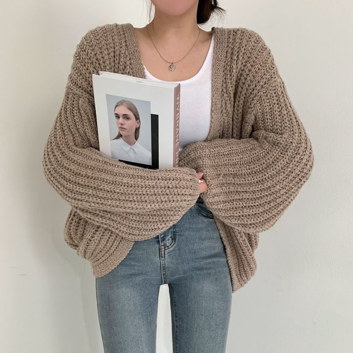 Frau in beiger Strickjacke, hält Buch, trägt weiße Bluse und Jeans, modisch, lässiger Stil.
