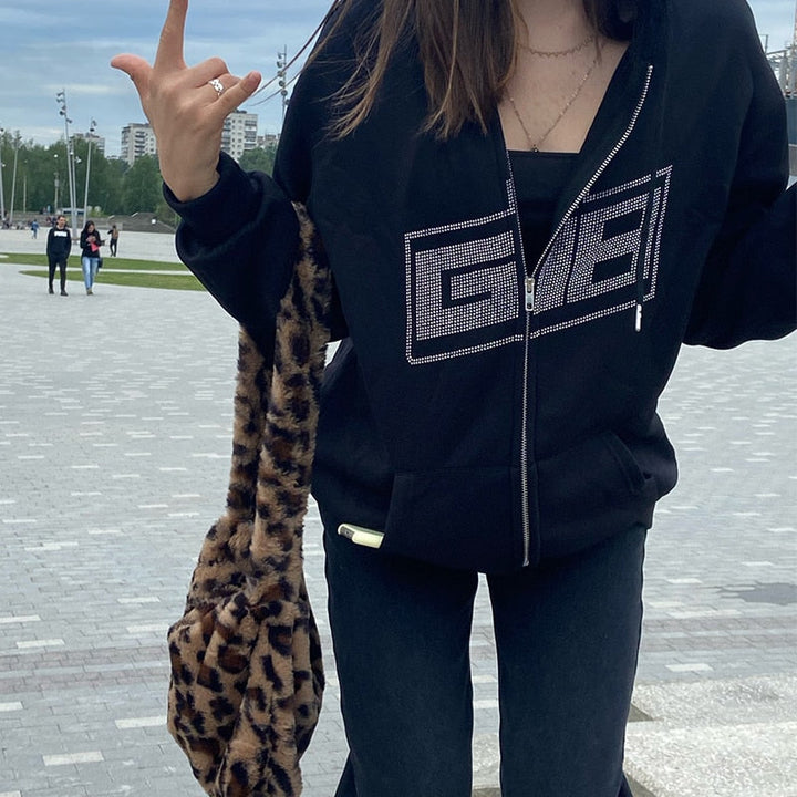 Frau in schwarzem Hoodie mit Glitzer-Logo, Jeans und Leopardenmuster-Tasche im Freien.