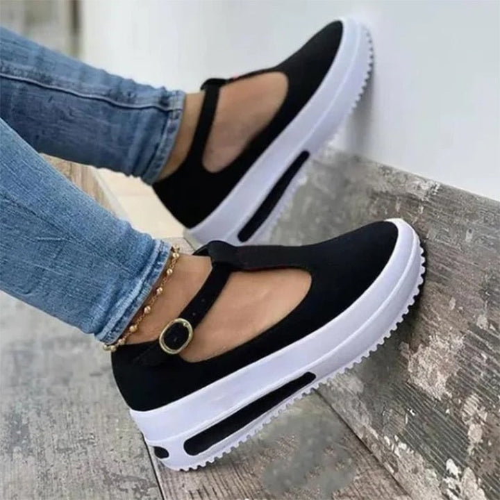 Schwarze Damen-Sneaker mit Plateau, T-Riemen und Schnalle, kombiniert mit blauen Jeans.