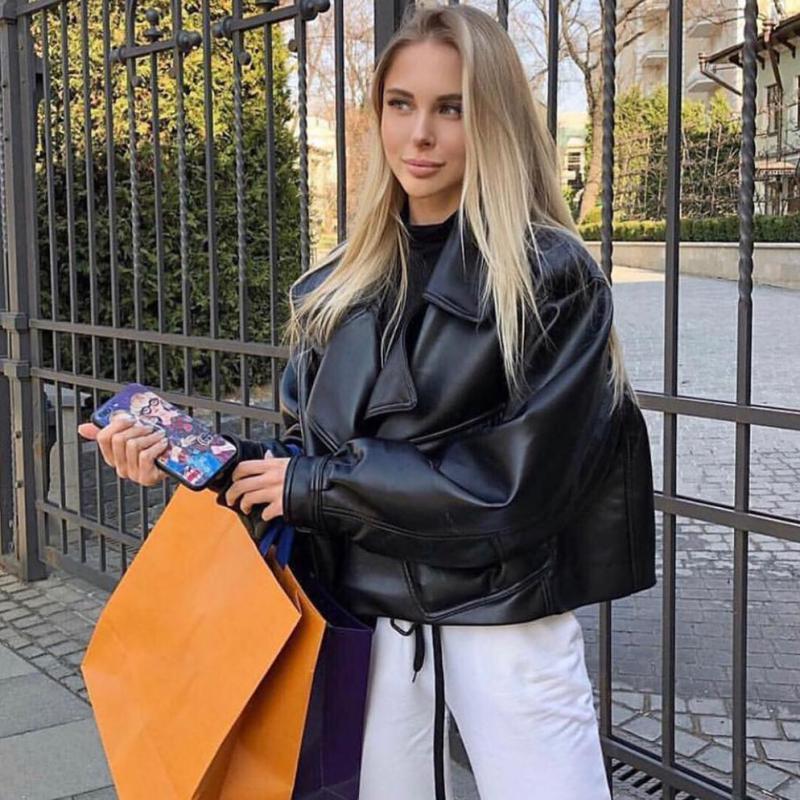 Blonde Frau in schwarzer Lederjacke mit Einkaufstaschen, Smartphone in der Hand, draußen.