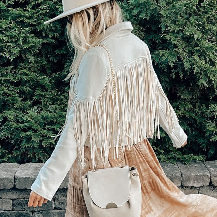 Frau mit weißer Fransenlederjacke und Hut, beige Tasche, vor grüner Hecke.