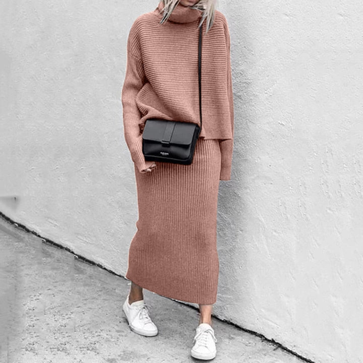 Frau in rosa Strickkleid mit schwarzer Umhängetasche und weißen Sneakers vor grauer Wand.