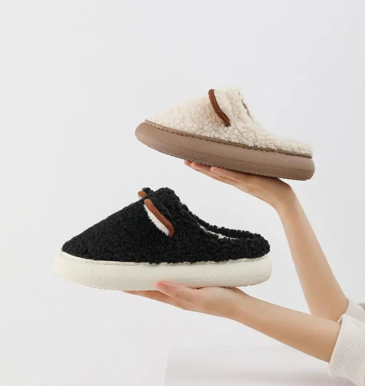Zwei flauschige Hausschuhe, schwarz und beige, mit rutschfester Sohle, in Händen gehalten.