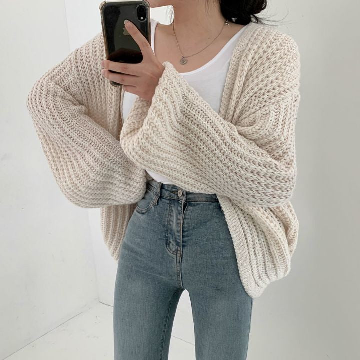 Frau in cremefarbenem Strickcardigan, weißem T-Shirt und blauer Jeans, Smartphone in der Hand.