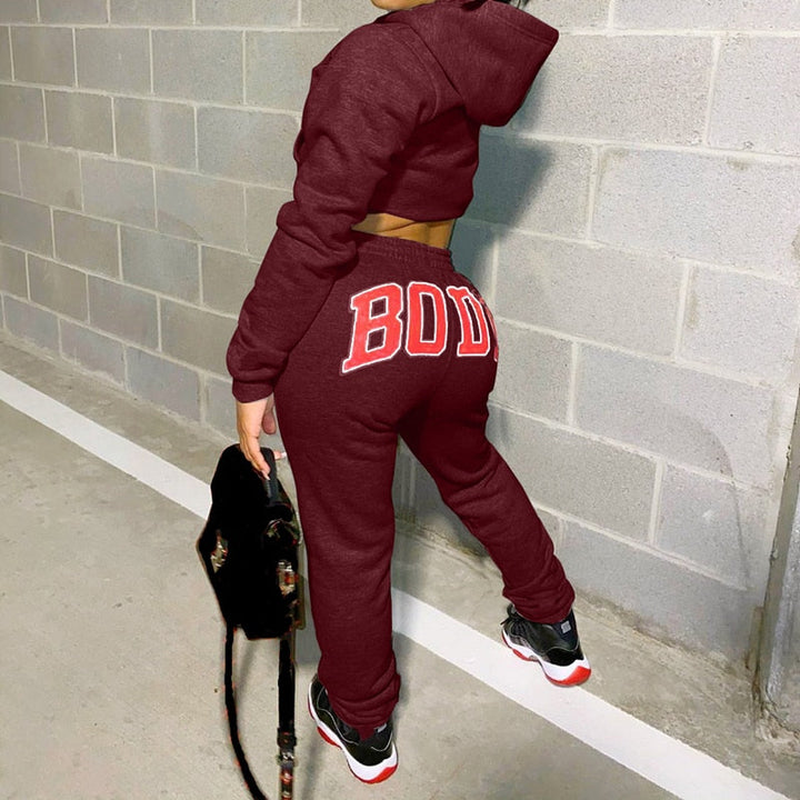 Frau in bordeauxfarbenem Jogginganzug mit "BODY"-Aufdruck, schwarze Sneaker, graue Wand.