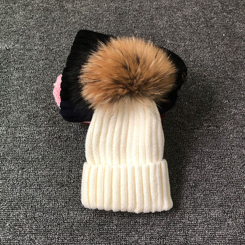 Weiße Strickmütze mit braunem Kunstfellbommel auf grauem Hintergrund.