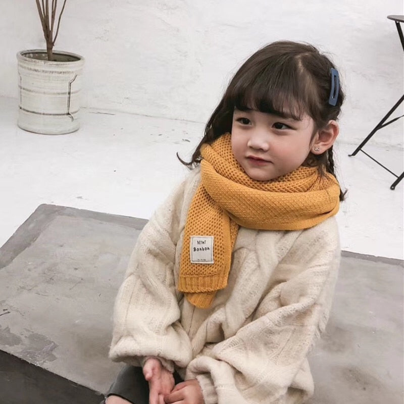 Mädchen mit gelbem Schal und weißem Pullover, sitzt auf grauer Bank, modisch, Winterkleidung.