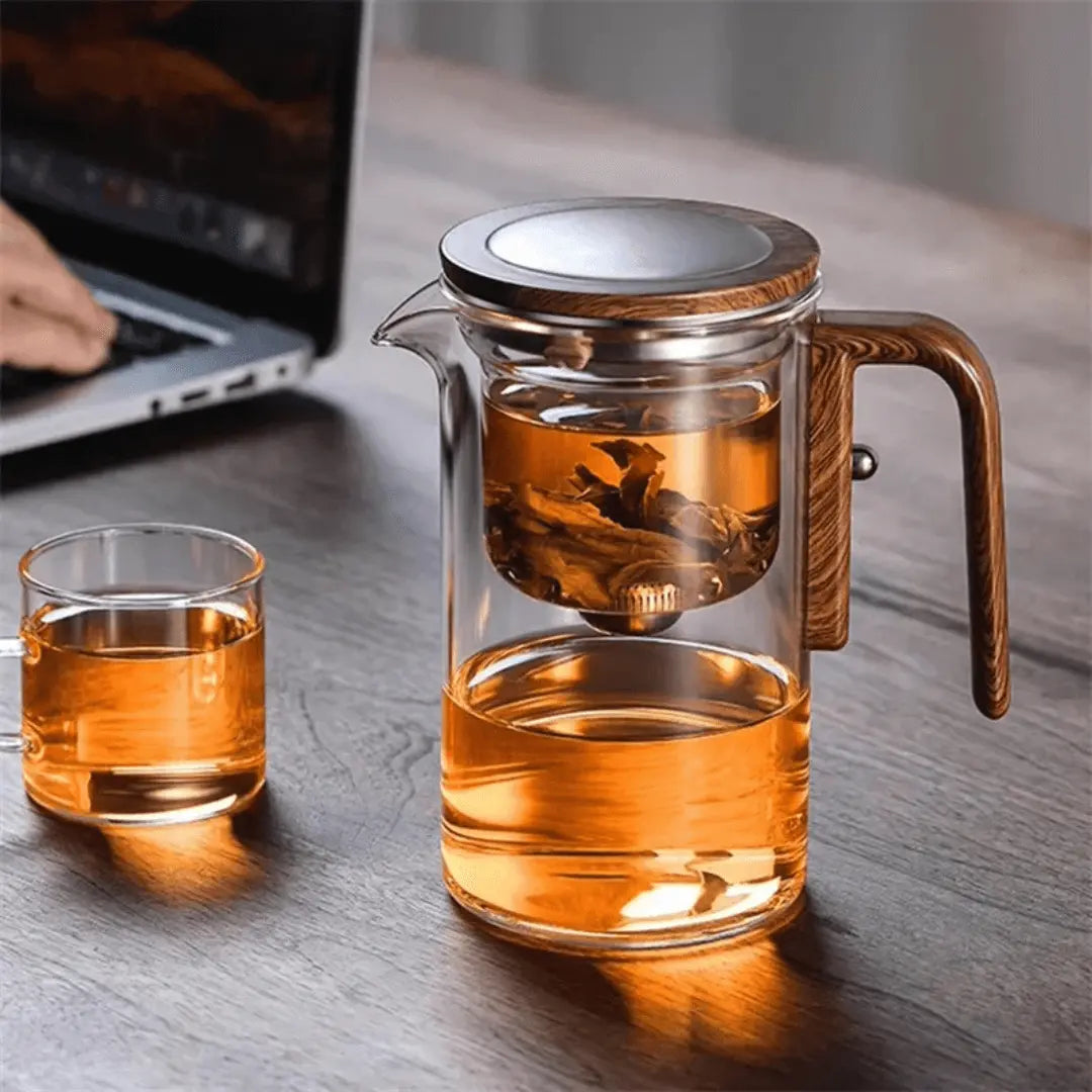 Glas-Teekanne mit Holzgriff und Sieb, gefüllt mit Tee, auf Holztisch neben Laptop.