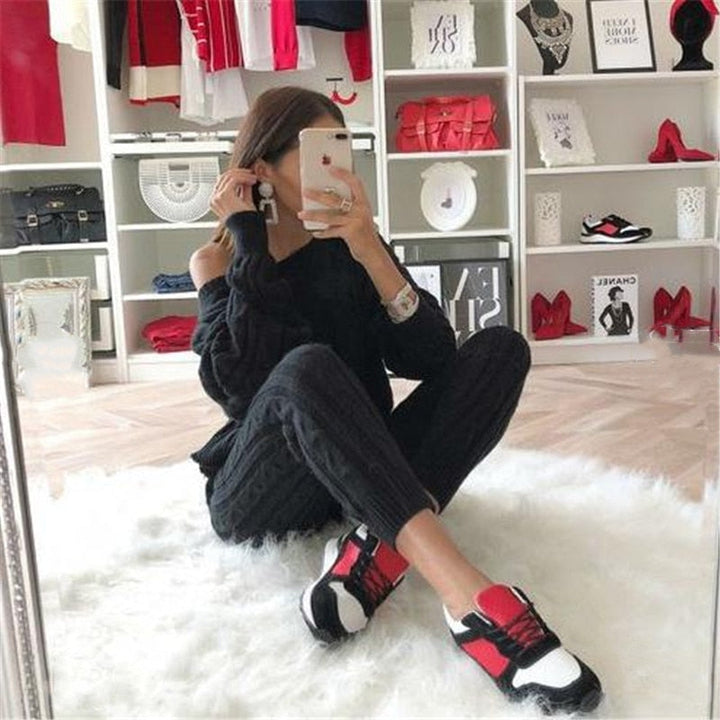 Frau in schwarzem Outfit mit rot-weißen Sneakers, sitzt auf weißem Teppich, modische Kleidung.