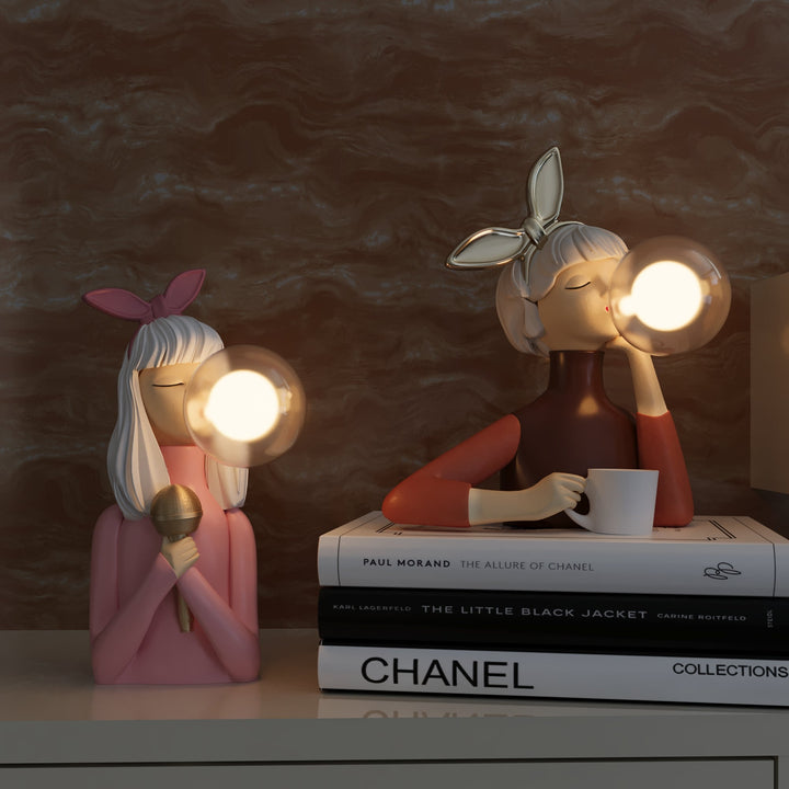 Zwei dekorative Tischlampen in Form von Frauenfiguren mit Kaugummi, rosa und braun, auf Bücherstapel.