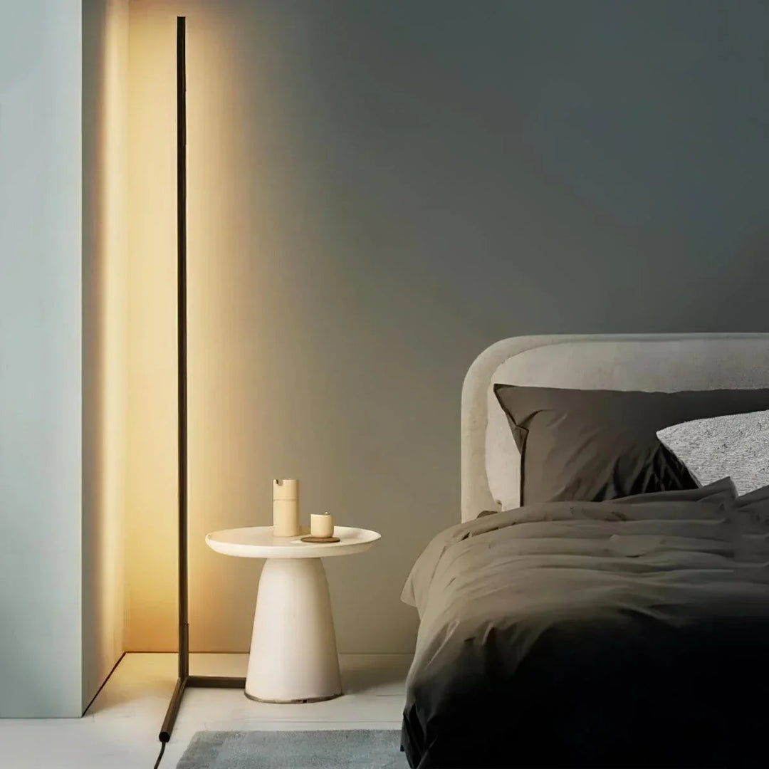 Rozsvícená tenká lampa v ložnici vedle bílého odkládacího stolku a postele s tmavým povlečením 1