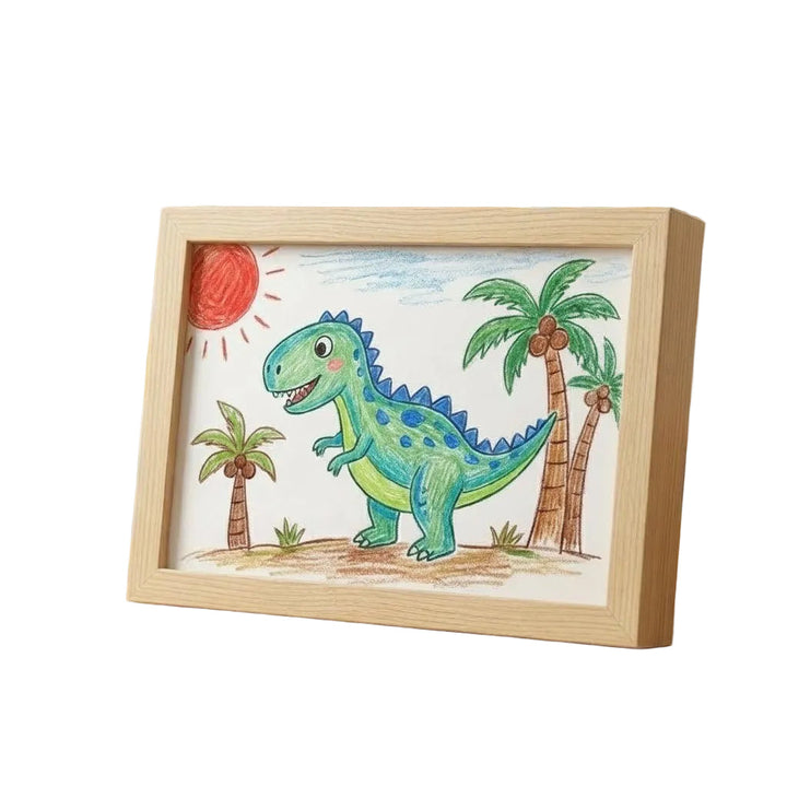 Světlý dřevěný rám s dětskou pastelkovou kresbou zeleného dinosaura mezi palmami a sluncem, vyfocený na bílém pozadí z boku. 1