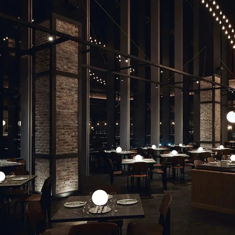 Prostorná tmavá restaurace s cihlovými pilíři, na jejíchž stolech září bílé světelné koule jako stolní osvětlení. 8