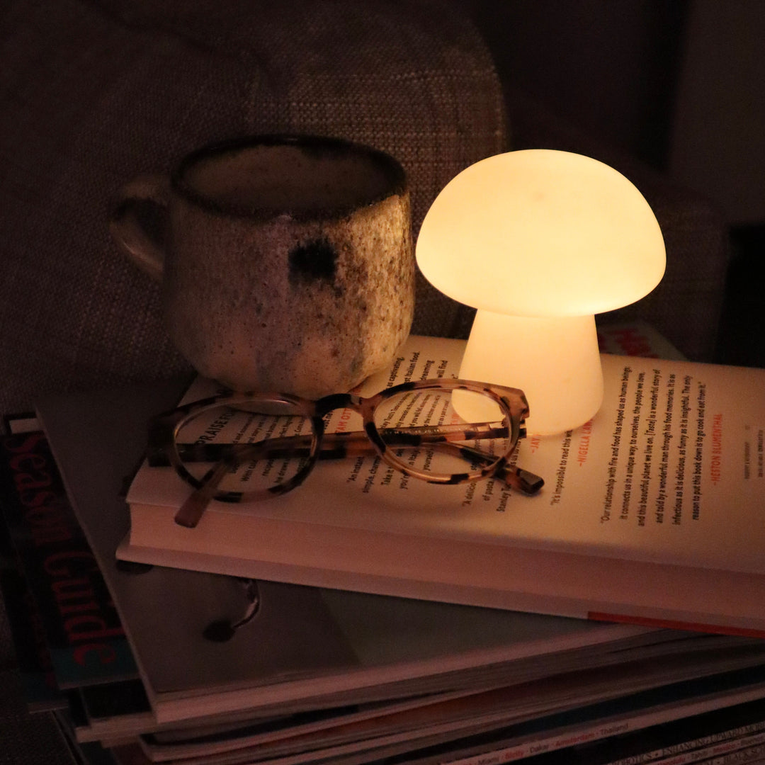 Keramikbecher, leuchtende Pilzlampe, Brille auf Buch, gemütliche Atmosphäre, Wohnzimmer-Dekor.