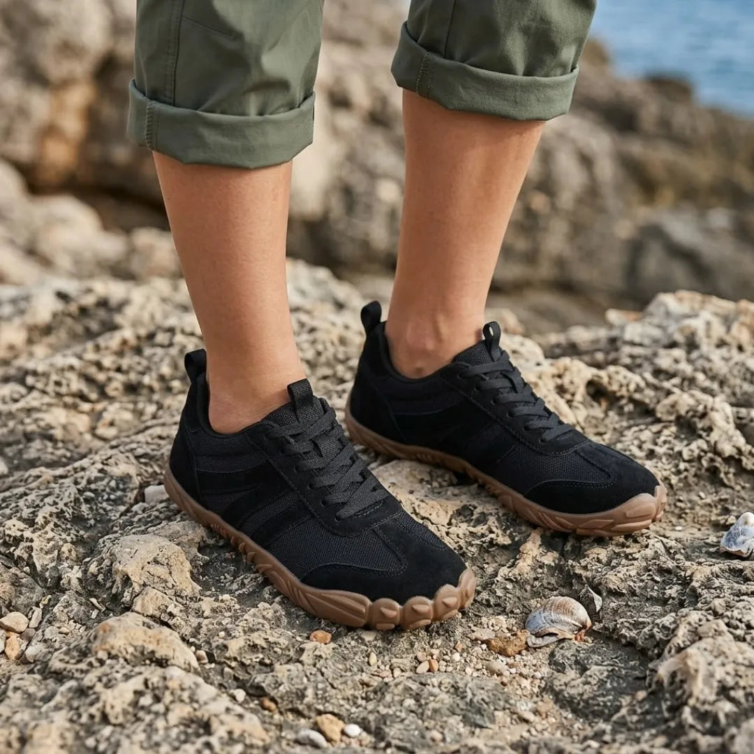 Černé barefoot tenisky na nohou osoby v zelených kalhotách s ohrnutými nohavicemi, stojící na skalnatém povrchu u moře. 20