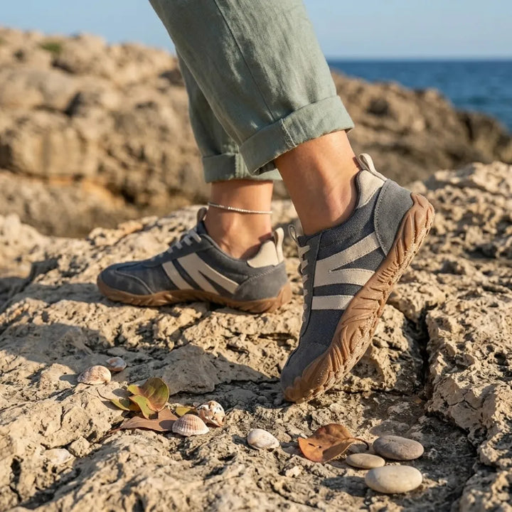 Šedé barefoot tenisky s béžovými pruhy na nohou ženy v zelených kalhotách, jak kráčí po skalnatém pobřeží u moře. 2