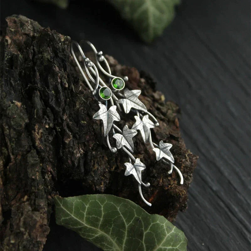 Silberne Ohrringe mit Efeublatt-Design und grünen Steinen auf Holzuntergrund.