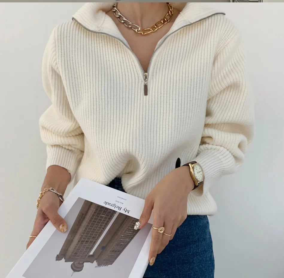 Frau in cremefarbenem Strickpullover mit Reißverschluss, goldener Schmuck, hält Buch.
