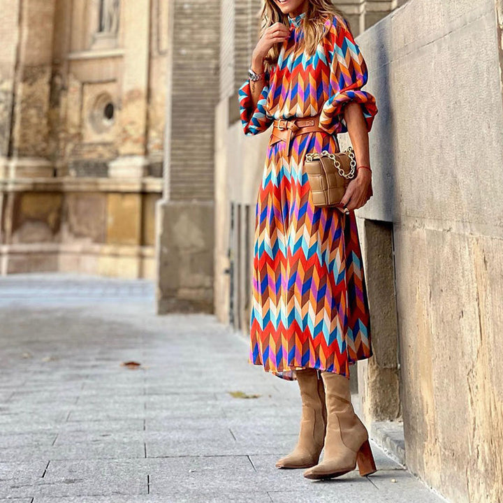 Frau in bunter Midi-Kleid mit geometrischem Muster, braunen Stiefeln und Tasche auf Straße.