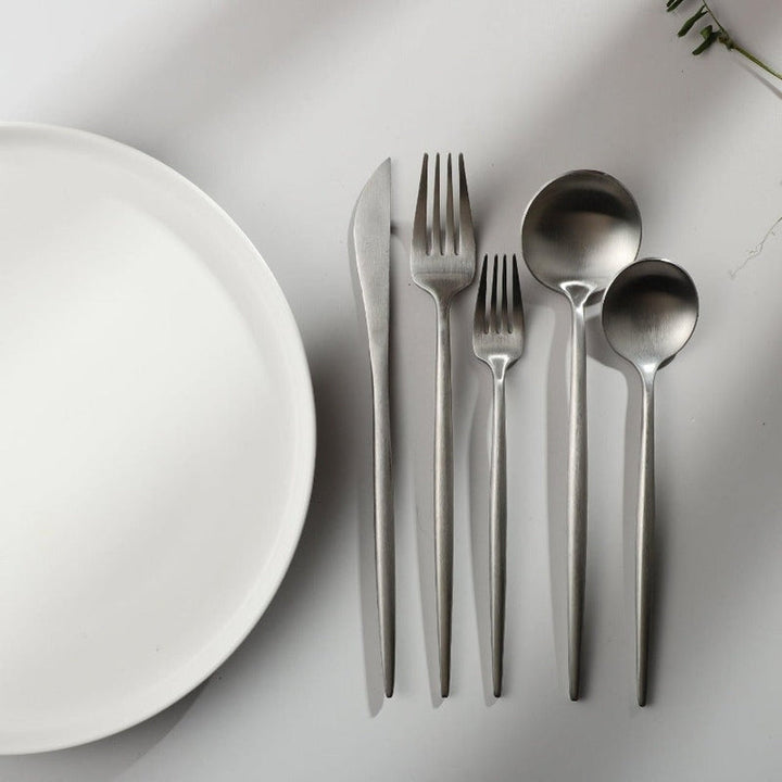Minimalistisches Besteckset aus Edelstahl neben weißem Teller auf grauem Hintergrund.