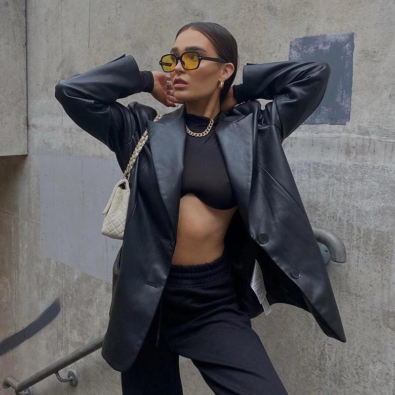 Frau in schwarzer Lederjacke, gelbe Sonnenbrille, schwarze Hose, weiße Tasche, urbane Umgebung.