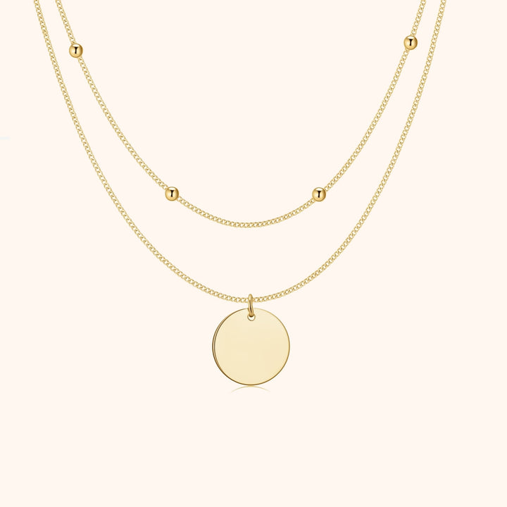 Zweireihige goldene Halskette mit rundem Anhänger und kleinen Buchstabenperlen.
