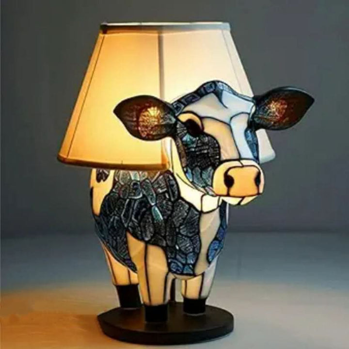 Lampa s krémovým stínidlem na stolní podstavě, jejíž základ tvoří vitrážová kráva v modrobílých barvách. 12