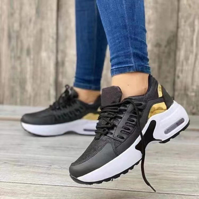 Frau trägt schwarze Sneaker mit goldenen Akzenten und weißer Sohle auf Holzboden.
