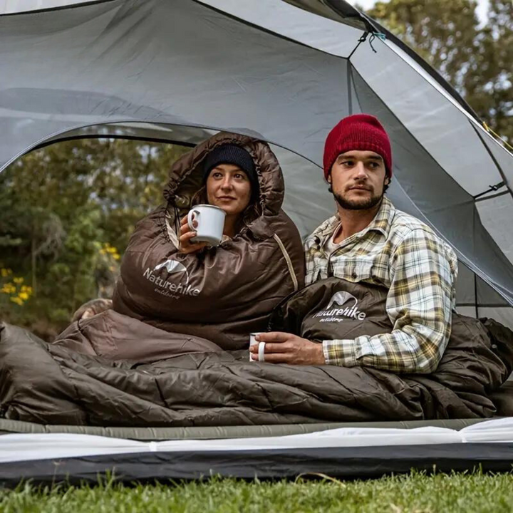 Zwei Camper in braunen Naturehike-Schlafsäcken im Zelt, trinken Kaffee, Outdoor-Abenteuer.