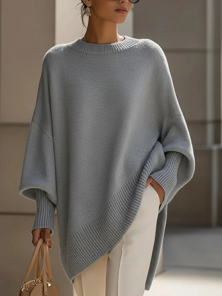 Šedý oversized svetr s asymetrickým žebrovaným lemem a širokými rukávy na modelce s béžovou kabelkou. 9