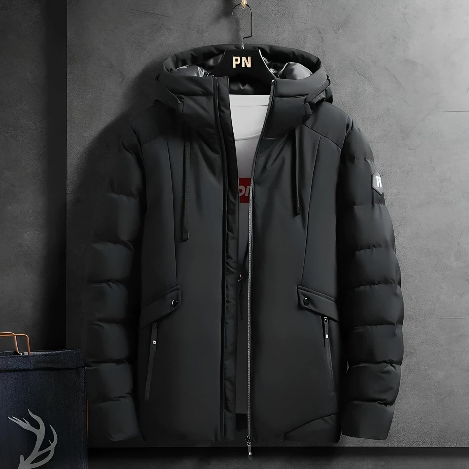 Schwarze Winterjacke mit Kapuze, Reißverschlüssen und Taschen, hängend an grauer Wand.