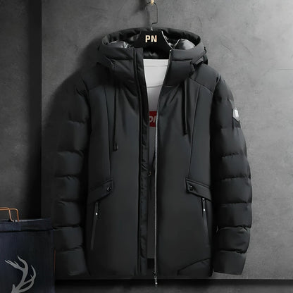 Schwarze Winterjacke mit Kapuze, Reißverschlüssen und Taschen, hängend an grauer Wand.