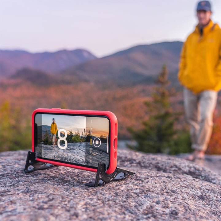 Rotes Smartphone im PocketTripod auf Felsen, Herbstlandschaft, Mann im Hintergrund.