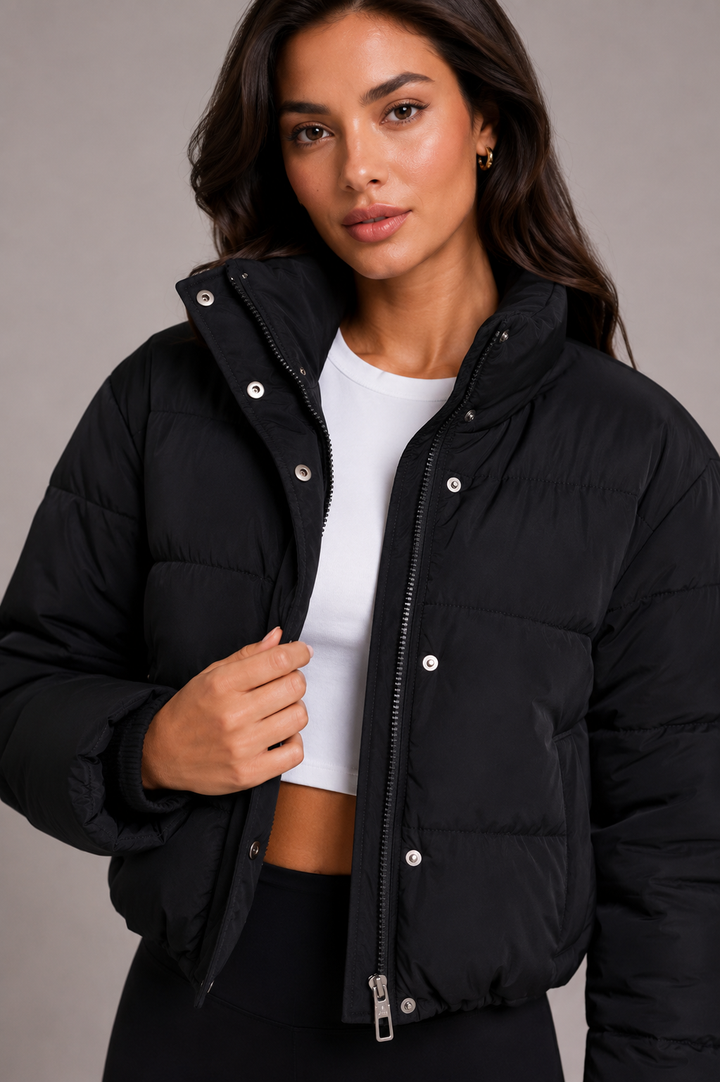 Detailní polodrážka modelky v černé prošívané bundě s vysokým límcem, rozepnutým zipem a patentkami, pod ní bílý top. 1