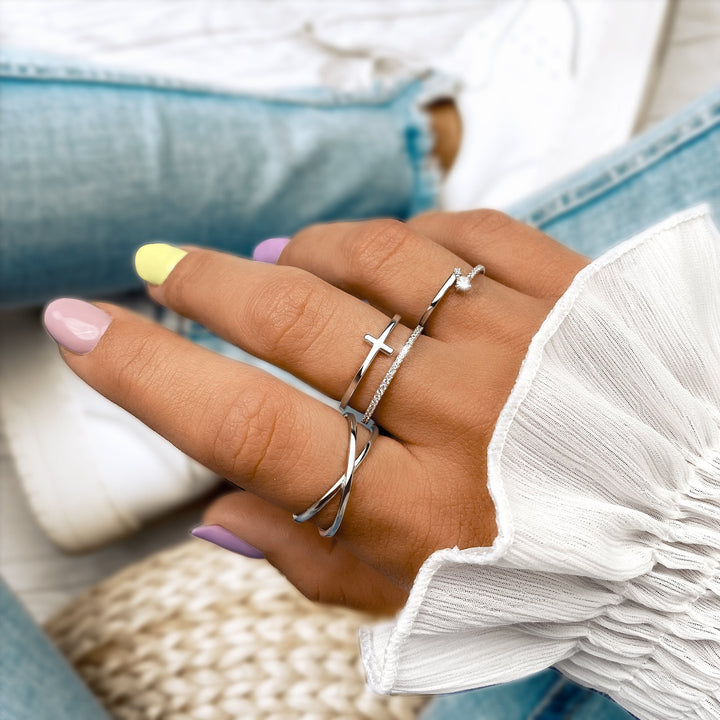 Damenhand mit silbernen Ringen, Kreuzdesign, pastellfarbene Nägel, weiße Bluse, Jeans.